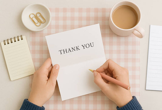 Top-down photo of a woman writing a thank you card at a pink gingham desk with a pen, notebook, coffee mug, and gold paperclips.