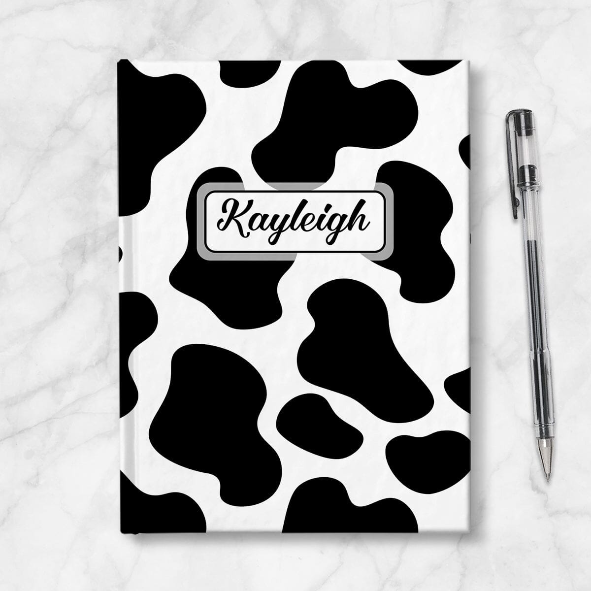 Personalized Cow Print Journal at Artistically Invited. A custom book illustrated with a bold and sleek black and white cow print pattern that wraps around the cover of the journal. Your personalized name is custom printed in a cute script font in black within a white rectangular design on the front of the book over the cow print background. Image shows the book next to a pen.