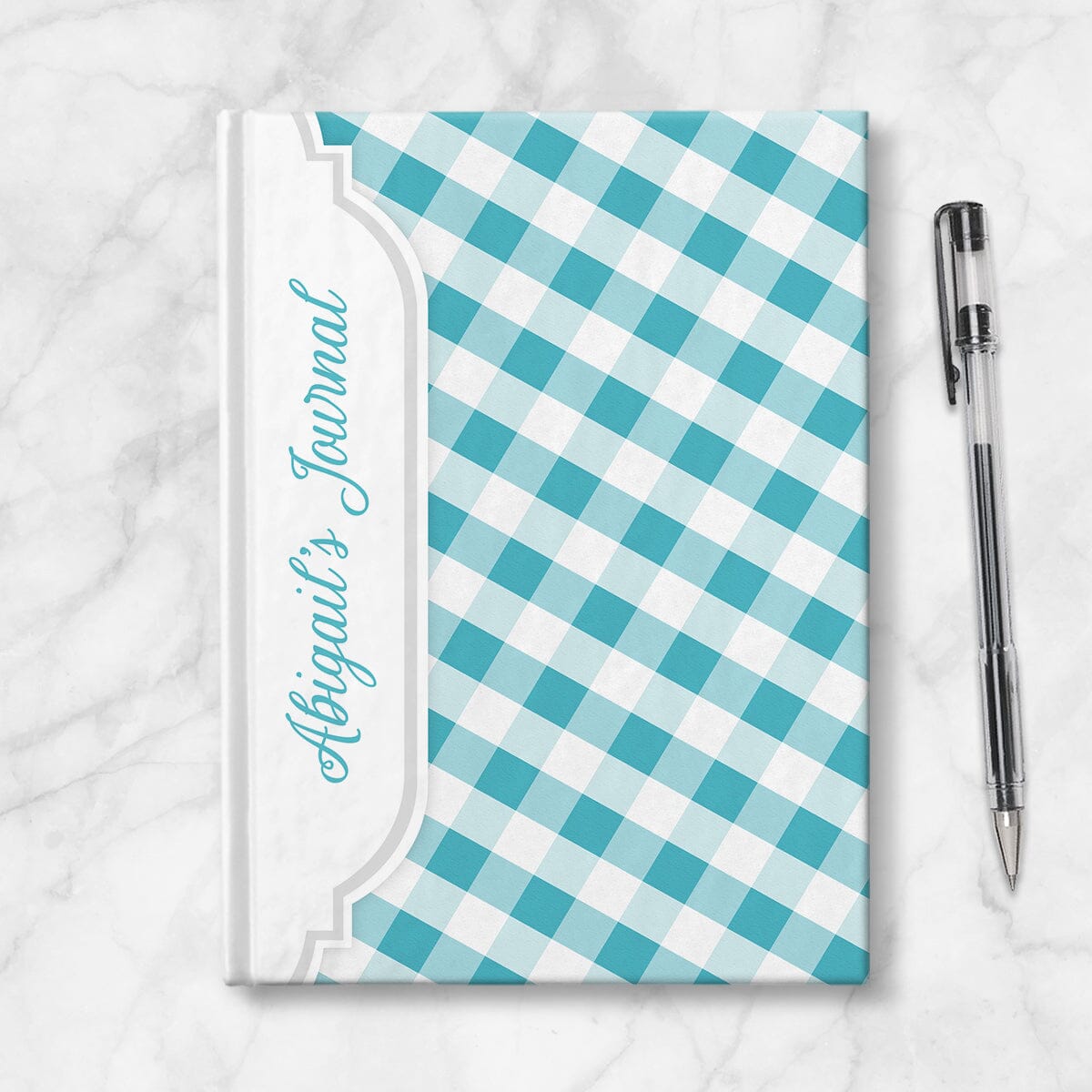 Personalized Turquoise Gingham Journal at Artistically Invited. Image shows the book on a countertop next to a pen.