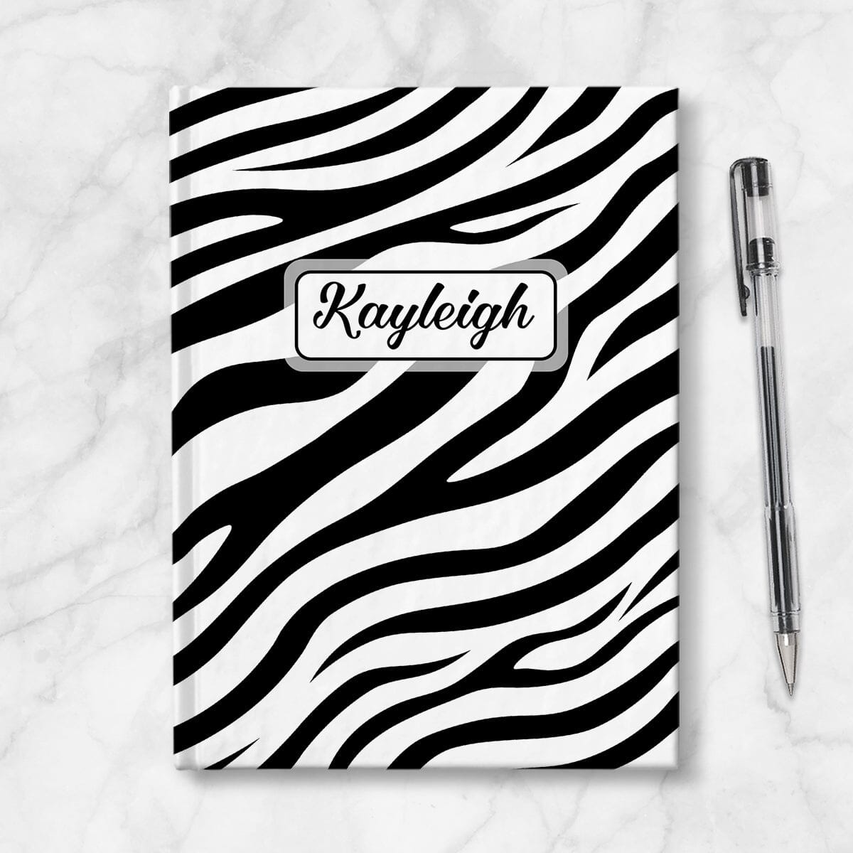 Personalized Zebra Print Journal at Artistically Invited. A custom book illustrated with a bold and sleek black and white zebra print pattern that wraps around the cover of the journal. Your personalized name is custom printed in a cute script font in black within a white rectangular design on the front of the book over the zebra print background. Image shows the journal next to a pen.