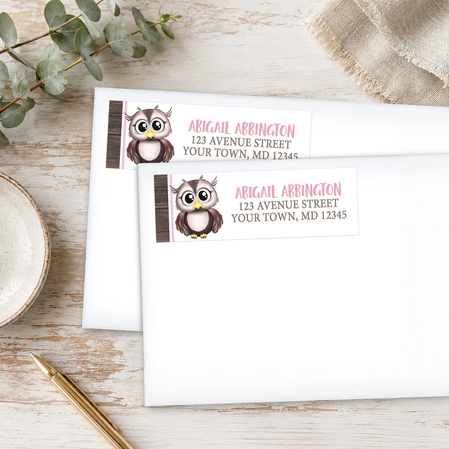 Address labels with owl design on a wooden surface with eucalyptus leaves and a pen.