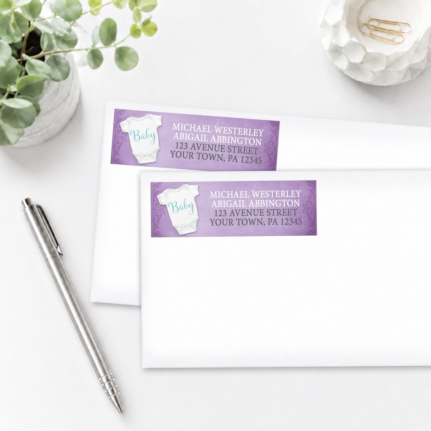 Baby Bodysuit on Purple Address Labels, shown on white enveloped, on a white surface with a plant and a pen.