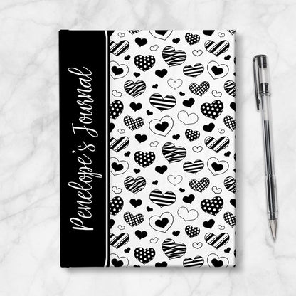 Personalized Black Heart Doodles Journal at Artistically Invited. A custom book featuring a pattern of hand-drawn hearts with different patterns and styles over a white background. Your personalized name is printed in a white handwriting font sideways on the front of the book over a black color block that wraps around the spine area. Image shows the book next to a pen.