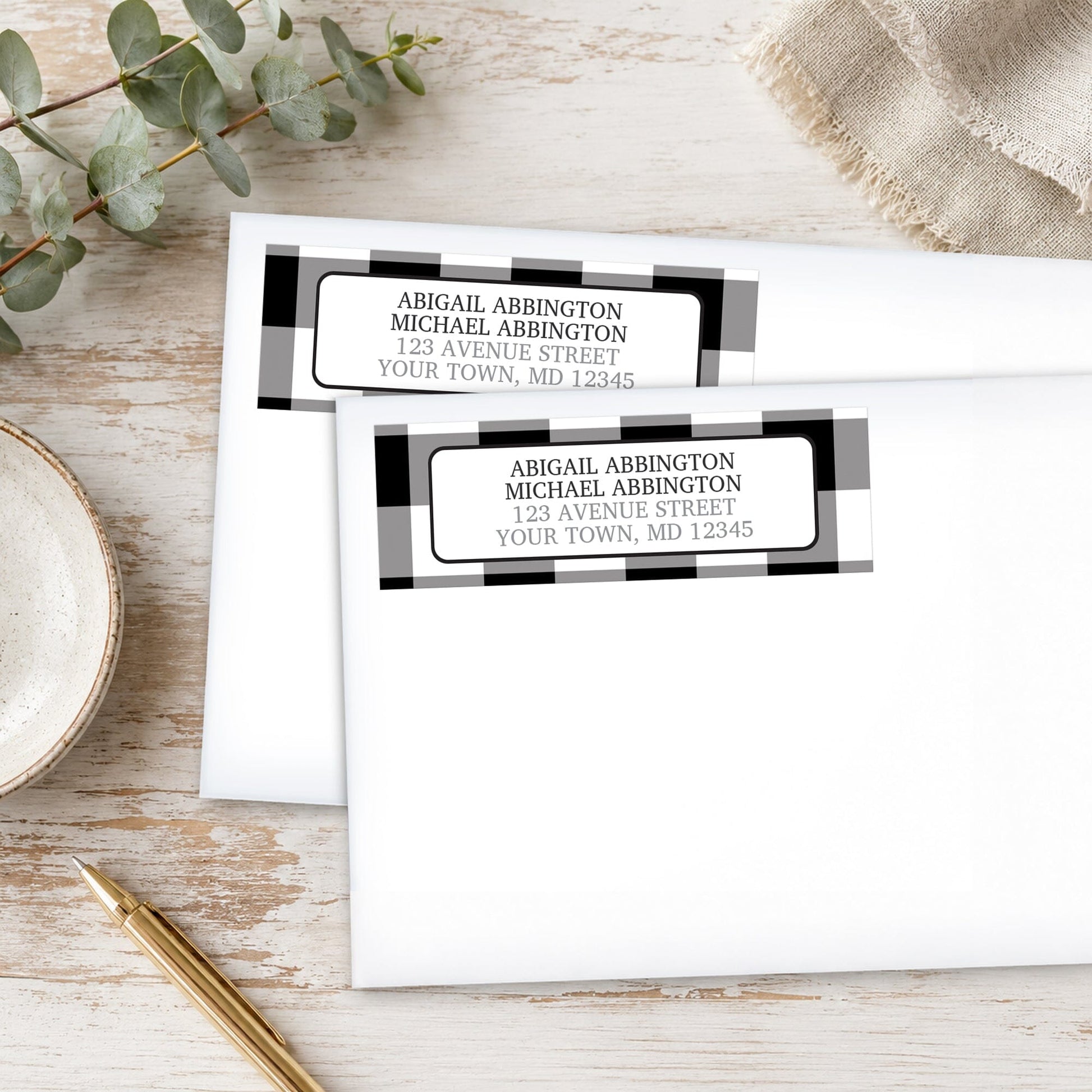Black and White Buffalo Plaid Address Labels, with check pattern borders, shown on white envelopes, with a gold pen and eucalyptus branch on a wooden surface.
