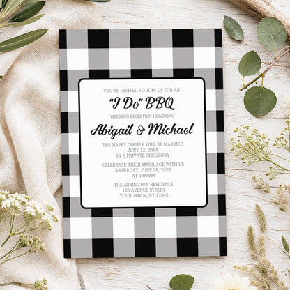 Black and white buffalo plaid I Do BBQ reception only wedding invitation with gray checks, black script lettering, and centered white wording panel, styled in a spring or summer-inspired flat lay with green leaves, white flowers, and a beige fabric background.