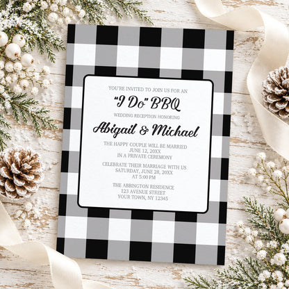Black and white buffalo plaid I Do BBQ reception only wedding invitation with gray checks, black script lettering, and centered white wording panel, styled in a flat-lay with pinecones, white berries, winter greenery, and a cream ribbon on a whitewashed wood background.