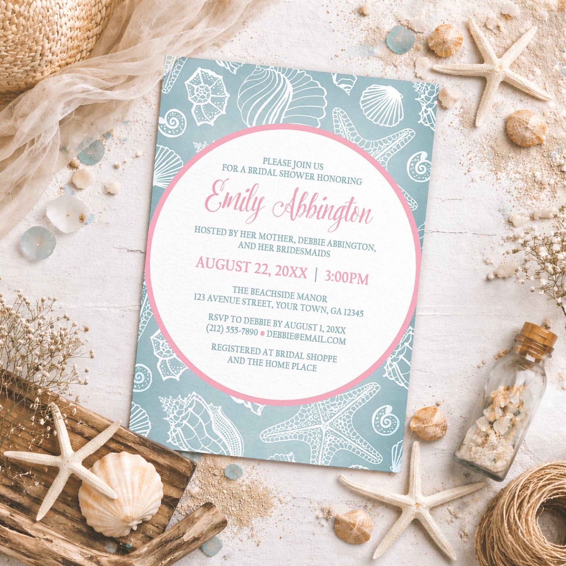Blue Seashell Pink Beach Bridal Shower Invitation shown on a beachy surface with sand and coastal decor.