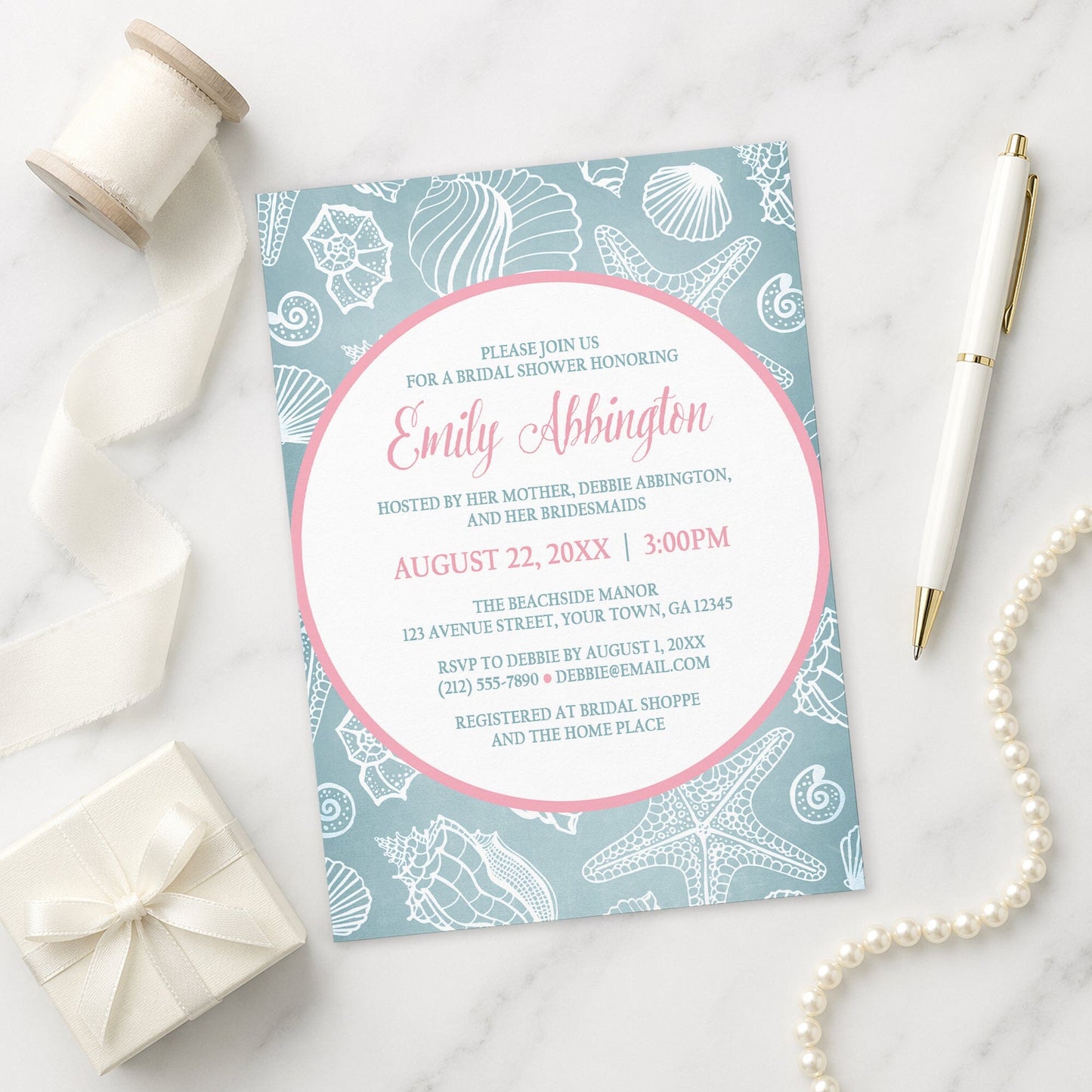 Blue Seashell Pink Beach Bridal Shower Invitation shown on a marble surface with a white gift box, pen, ribbon, and pearls.