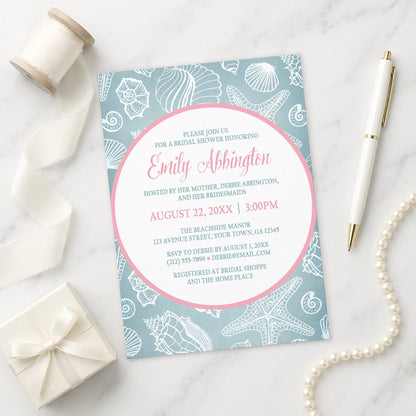 Blue Seashell Pink Beach Bridal Shower Invitation shown on a marble surface with a white gift box, pen, ribbon, and pearls.