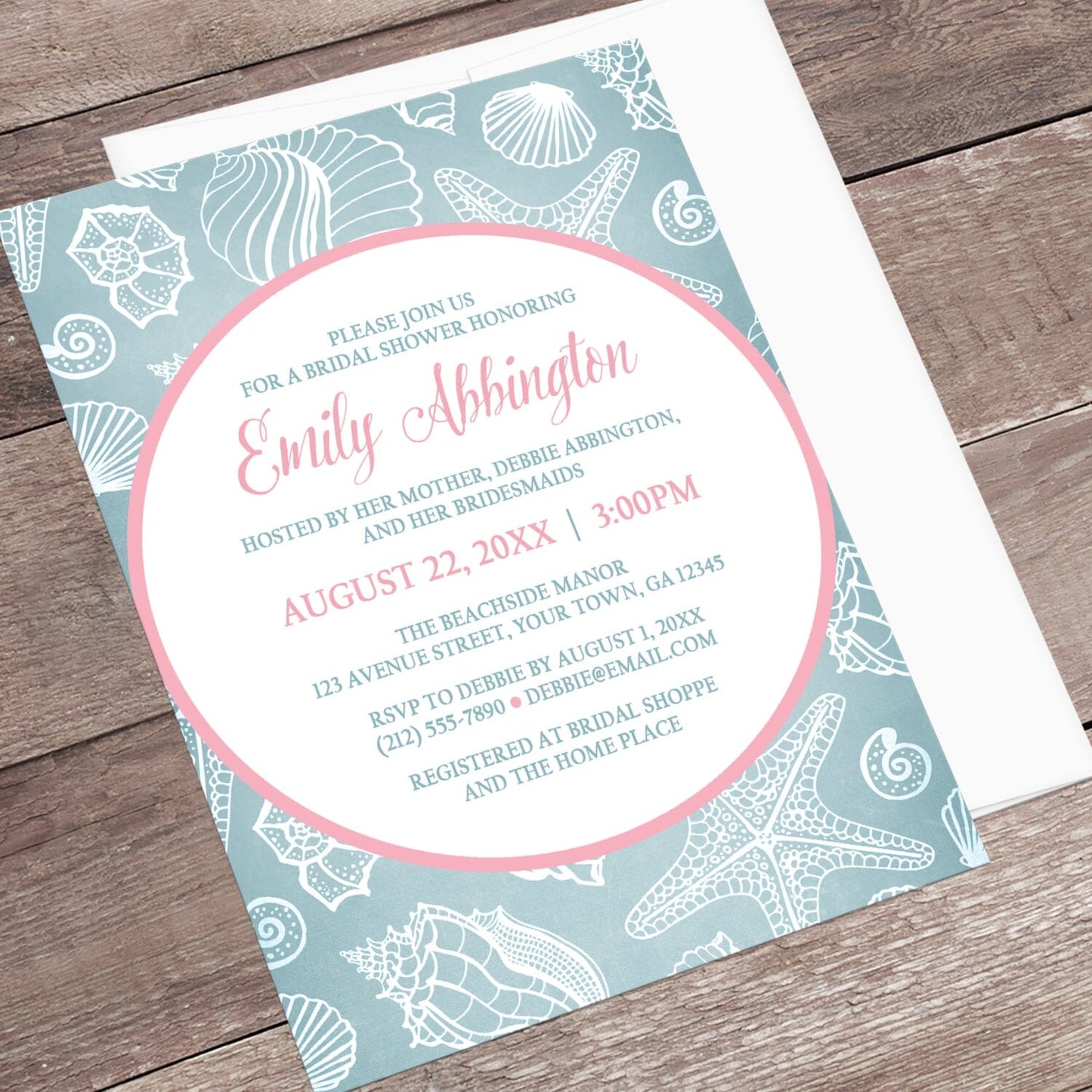 Blue Seashell Pink Beach Bridal Shower Invitation shown at an angle on a wooden surface with a white envelope.