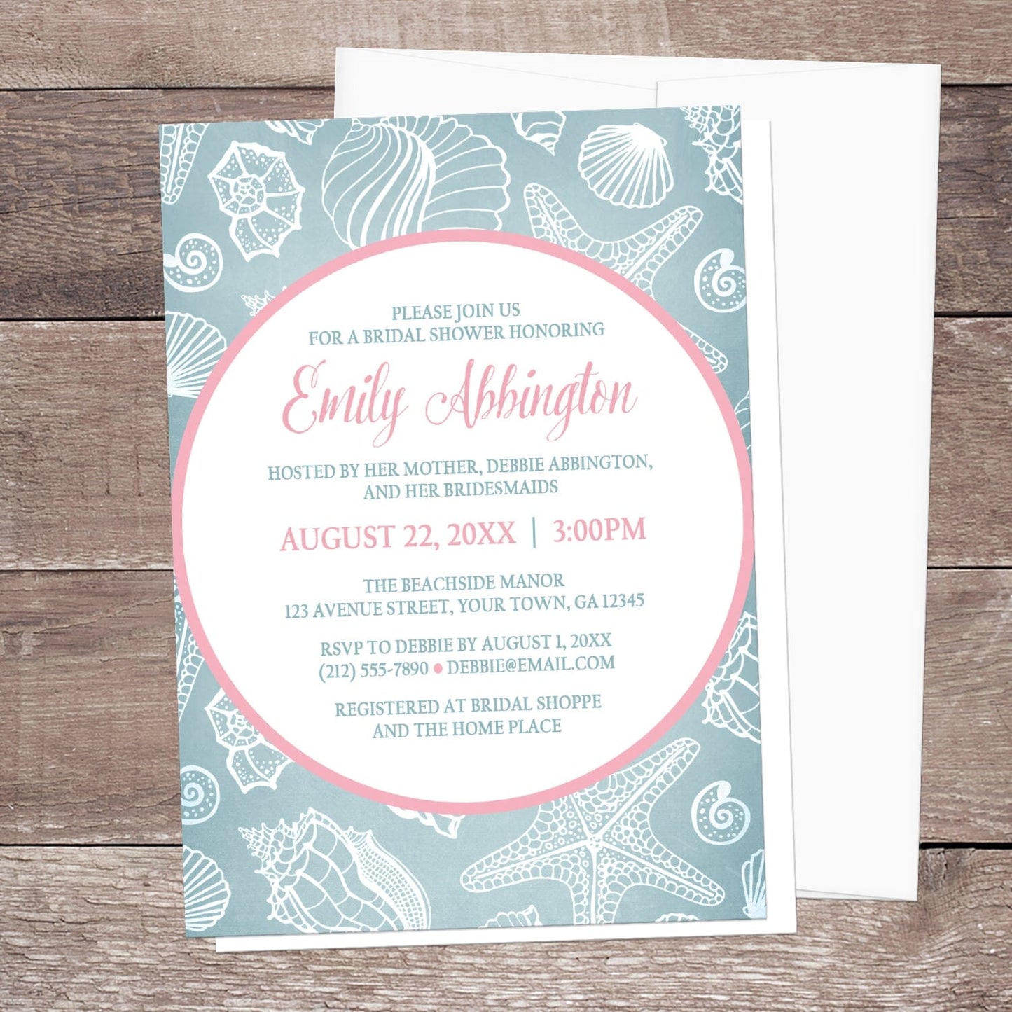 Blue Seashell Pink Beach Bridal Shower Invitation shown on a wooden surface with a white envelope.