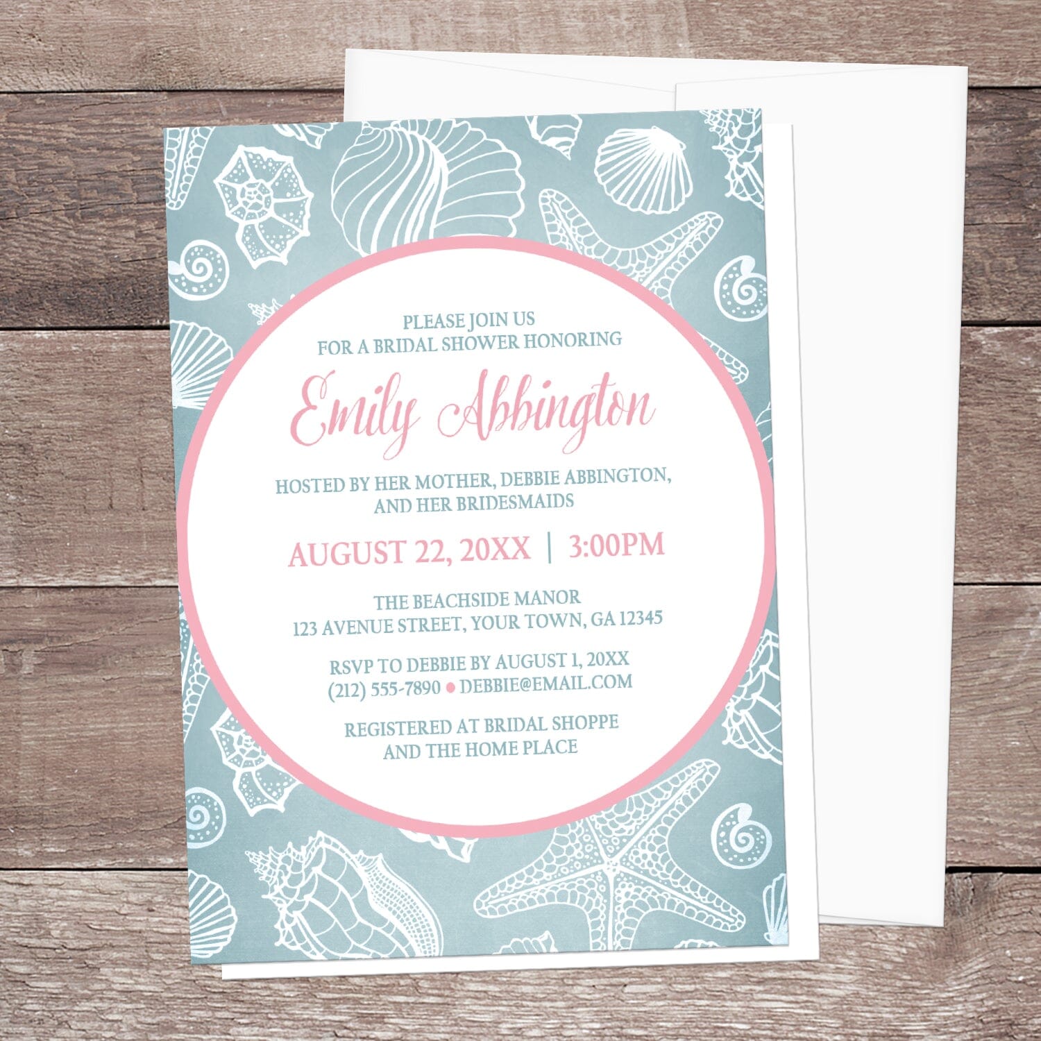 Blue Seashell Pink Beach Bridal Shower Invitation shown on a wooden surface with a white envelope.