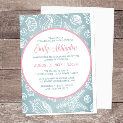 Blue Seashell Pink Beach Bridal Shower Invitation shown on a wooden surface with a white envelope.