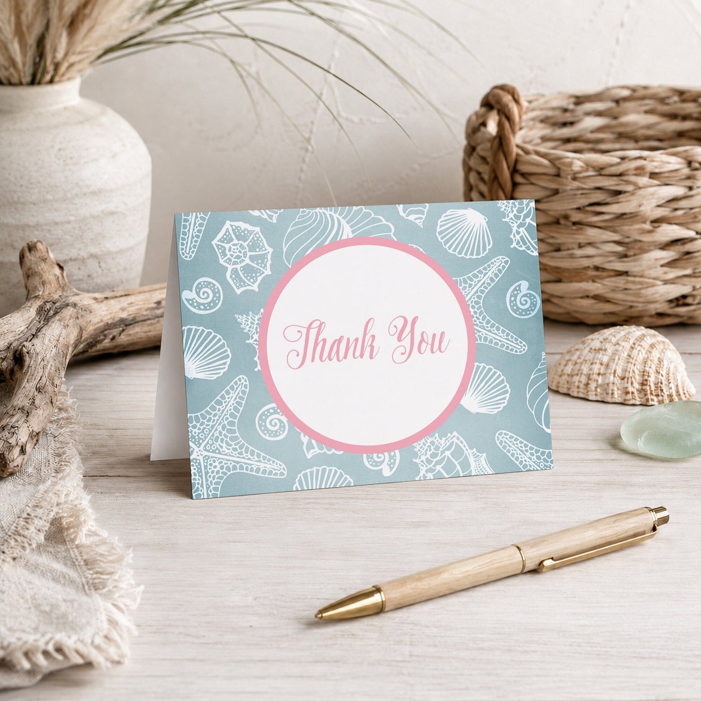 Blue Seashell Pink Beach Thank You Card shown on a beach house tabletop with coastal decor and a pen.
