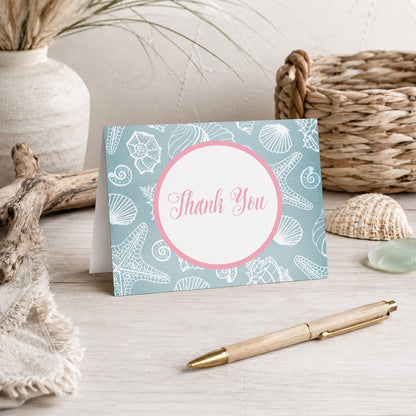 Blue Seashell Pink Beach Thank You Card shown on a beach house tabletop with coastal decor and a pen.