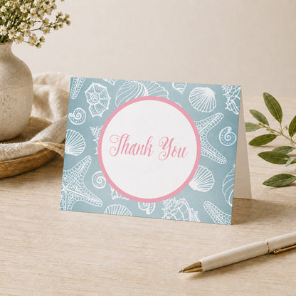 Blue Seashell Pink Beach Thank You Card shown on a desk with a vase, plants, and a pen.