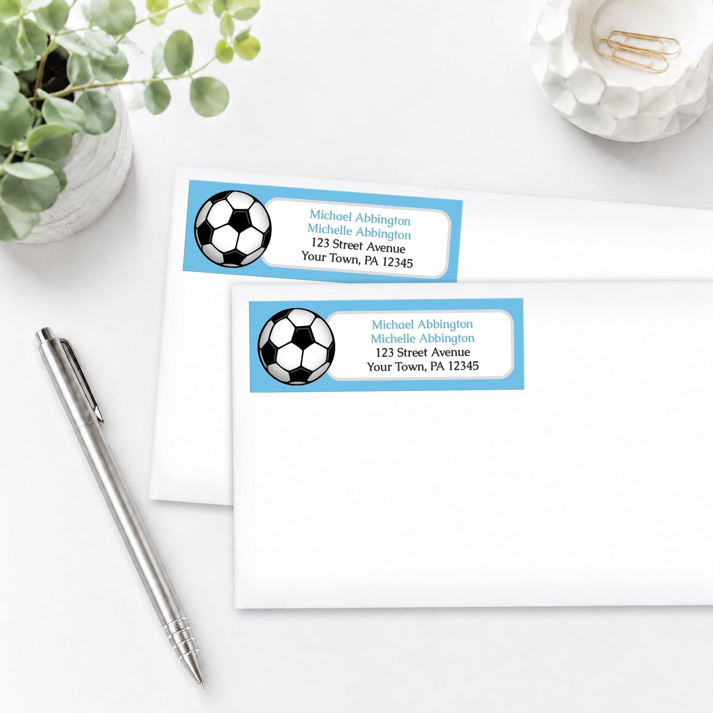 Blue Soccer Ball Address Labels on white envelopes, shown on a white surface with a plant and a pen.