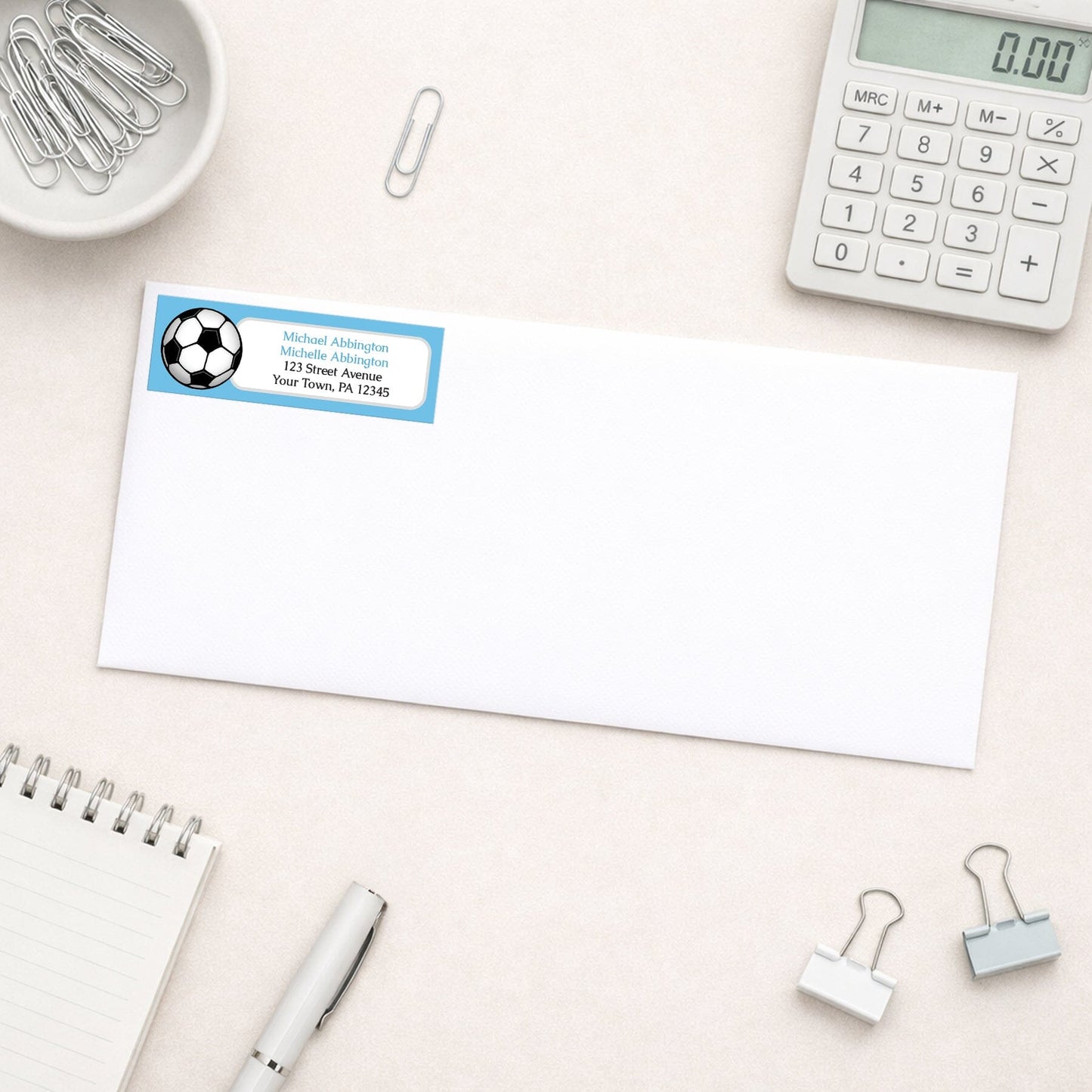 Blue Soccer Ball Return Address Label shown on a white envelope with everyday office supplies