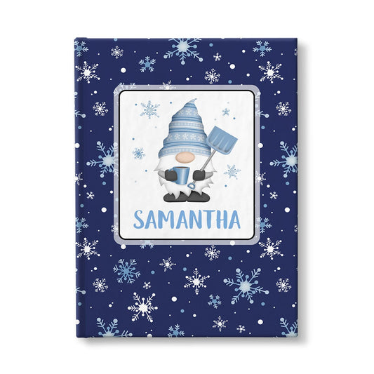 Personalized Blue Winter Gnome Journal at Artistically Invited. A custom book designed with an adorable gnome holding a hot beverage and a snow shovel with light blue snowflakes floating around him in a white rectangular design on the front of the book, with your name personalized in blue in a cute all capital letters font below the gnome. The background design that wraps around the cover of the book features a beautiful pattern whimsical snowflakes in white and blue over a navy blue background color.