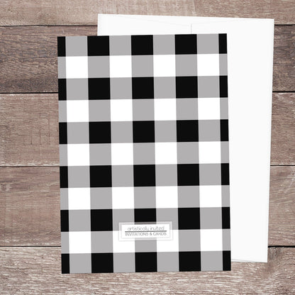 The back side of the Black and White Buffalo Plaid I Do BBQ Couples Shower Invitation, featuring the black and white check pattern, shown on a brown wood surface with a white envelope.