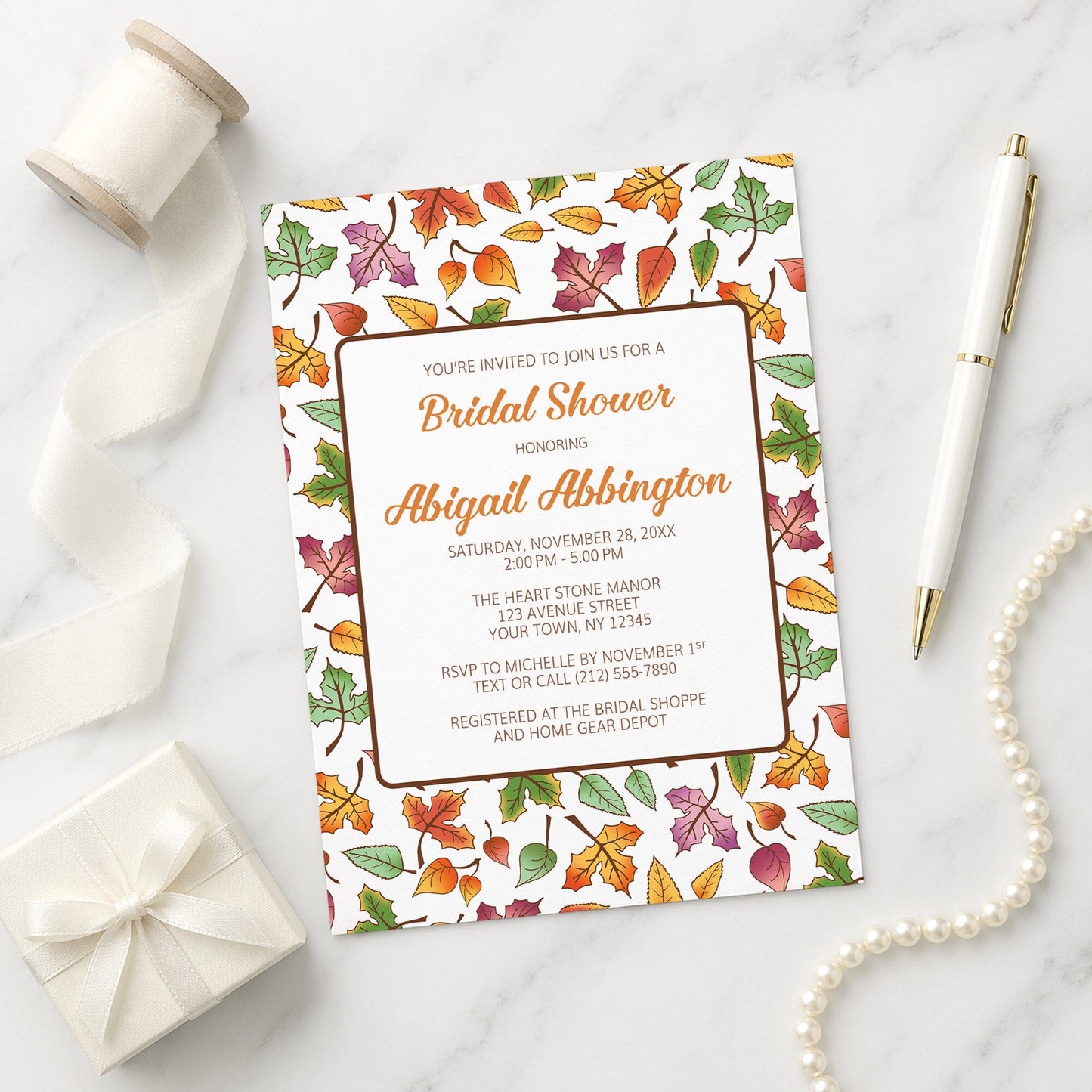 Bridal shower invitation with autumn leaf design on a marble surface with gift, pen, and necklace.