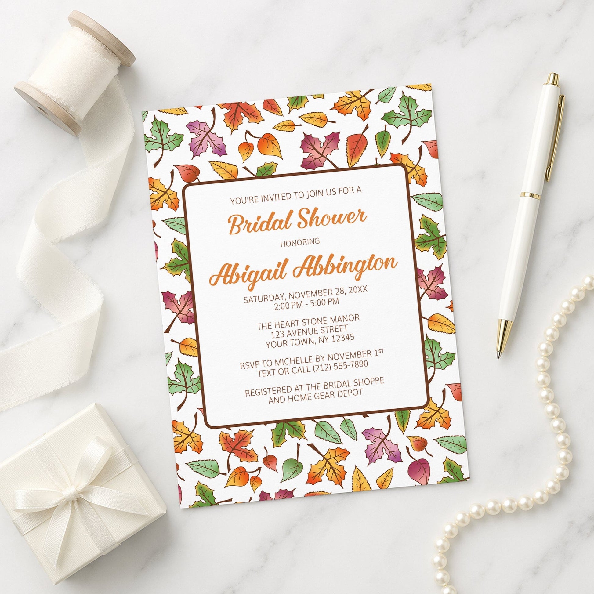 Bridal shower invitation with autumn leaf design on a marble surface with gift, pen, and necklace.