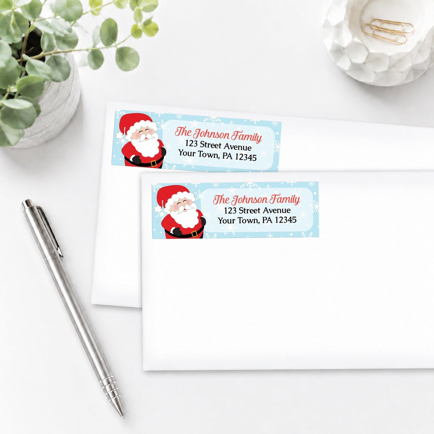 Cheerful Santa Address Labels with Santa Claus design, shown on white envelopes, on a white surface with a pen and plant.