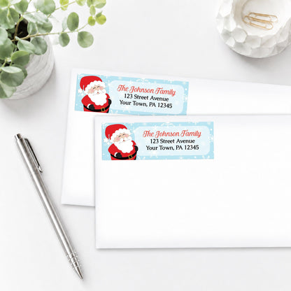 Cheerful Santa Address Labels with Santa Claus design, shown on white envelopes, on a white surface with a pen and plant.