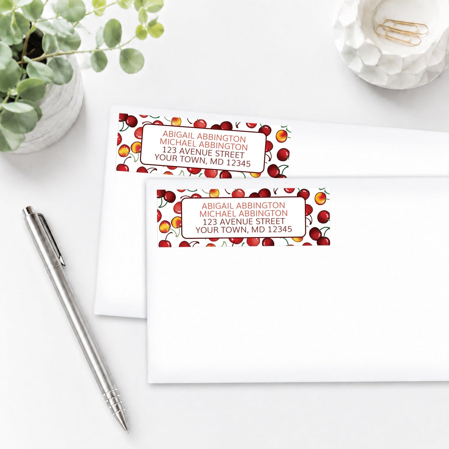Two Cherries Address Labels with cherry design, shown on white envelopes, on a white surface with a pen and plant.