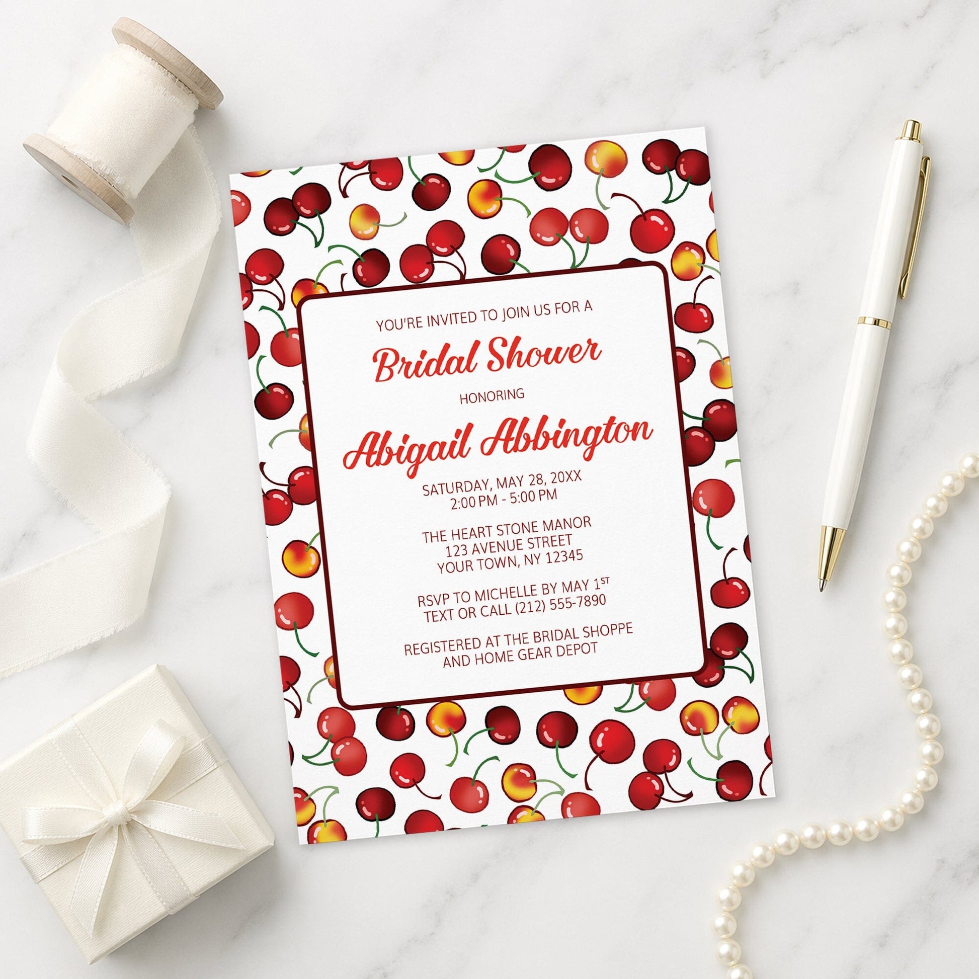 Bridal shower invitation with cherry design on a marble surface with ribbon, gift, and pen.
