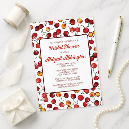 Bridal shower invitation with cherry design on a marble surface with ribbon, gift, and pen.