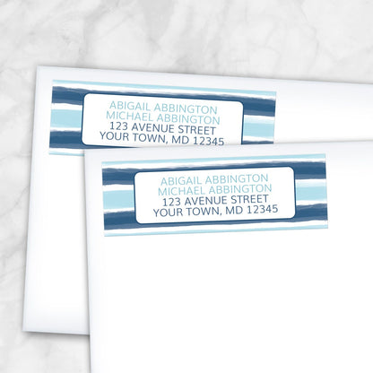 Coastal Paint Brush Strokes Address Labels with horizontal paint brush strokes-style stripes in navy and light blue and your return address custom printed in two shades of blue, shown on white envelopes on a marble surface.