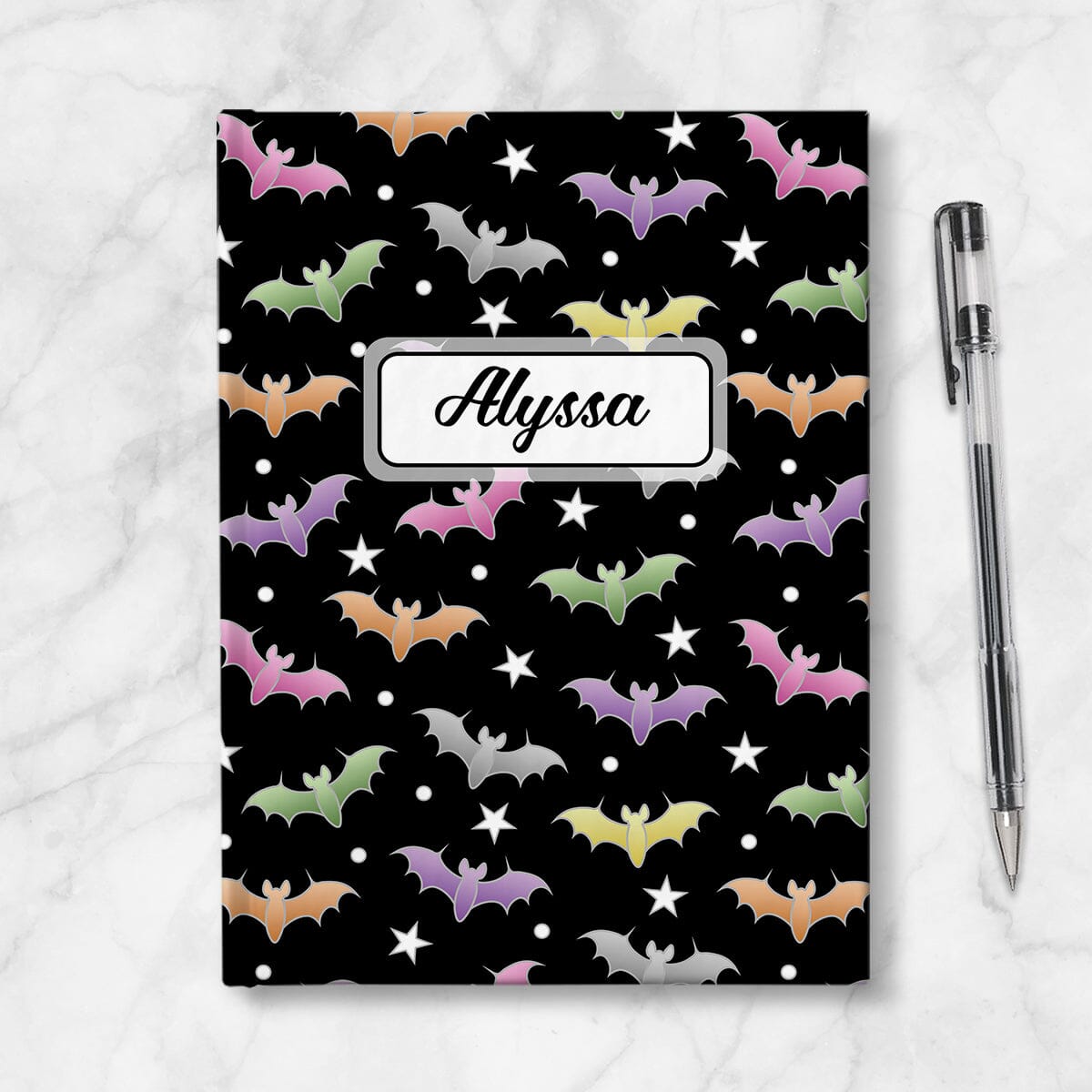 Colorful Bats Journal at Artistically Invited. A journal designed with a pattern of bats in different colors over a black background color that wraps around the book. Your personalized name is custom printed in a black script font in a white rectangular design over the bats pattern. Available with ruled lines or blank inside. Photo shows the journal next to a pen.
