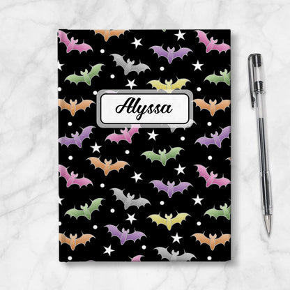 Colorful Bats Journal at Artistically Invited. A journal designed with a pattern of bats in different colors over a black background color that wraps around the book. Your personalized name is custom printed in a black script font in a white rectangular design over the bats pattern. Available with ruled lines or blank inside. Photo shows the journal next to a pen.