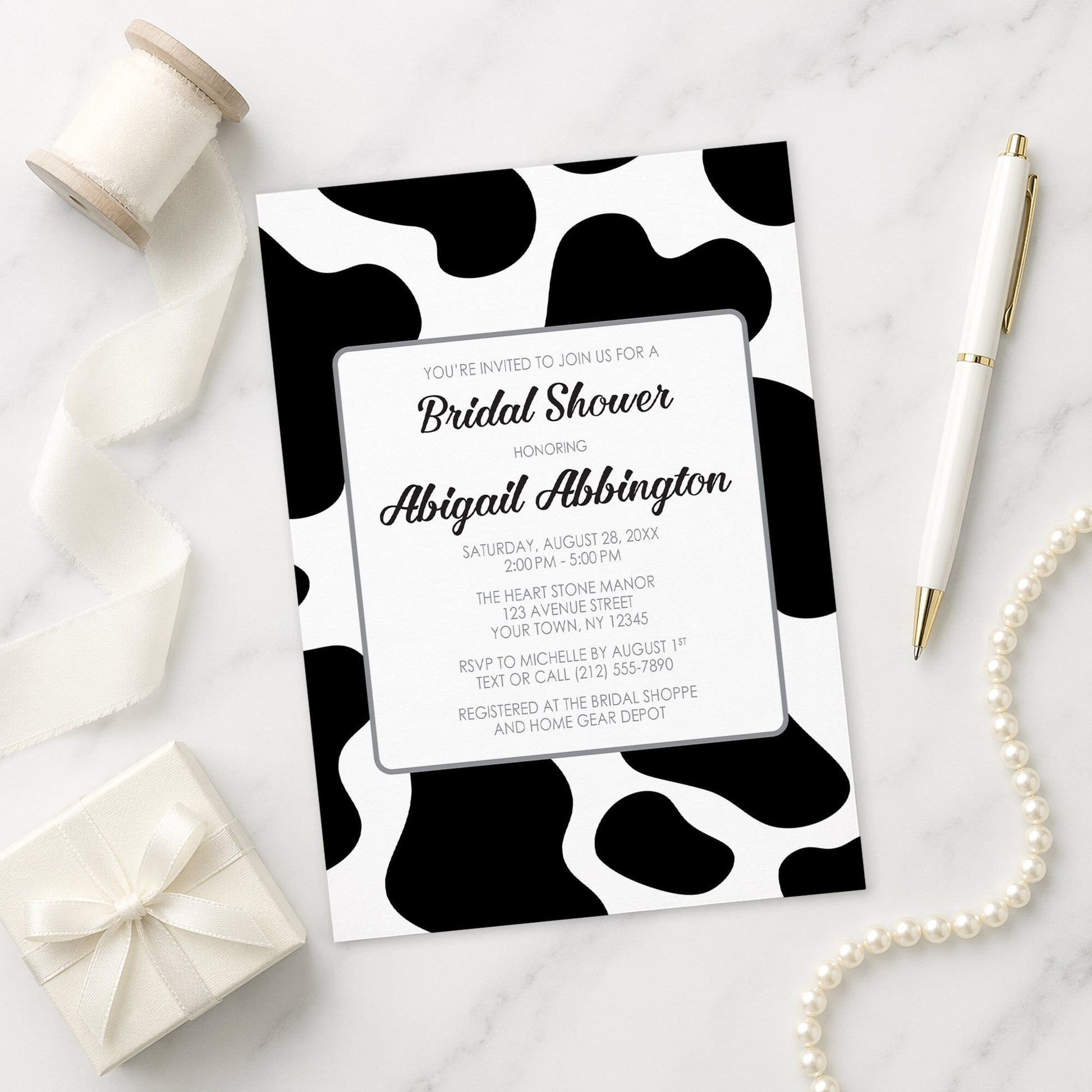 Cow Print Bridal Shower Invitation on a marble surface with gift, pen, and pearls.