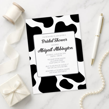 Cow Print Bridal Shower Invitation on a marble surface with gift, pen, and pearls.