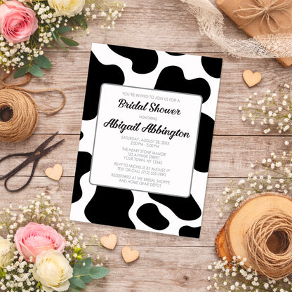Cow Print Bridal Shower Invitation on a rustic wooden surface with flowers, lace, and twine.