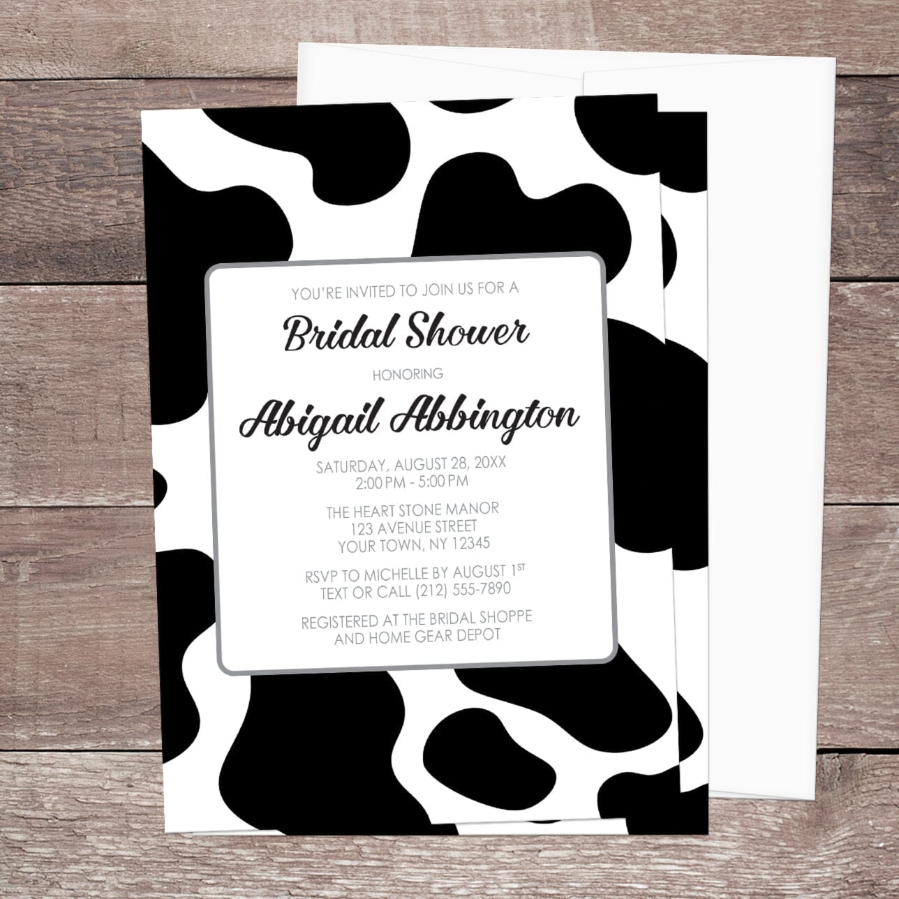 Bridal shower invitation with black and white cow spots pattern, displayed with a white envelope on a wooden surface