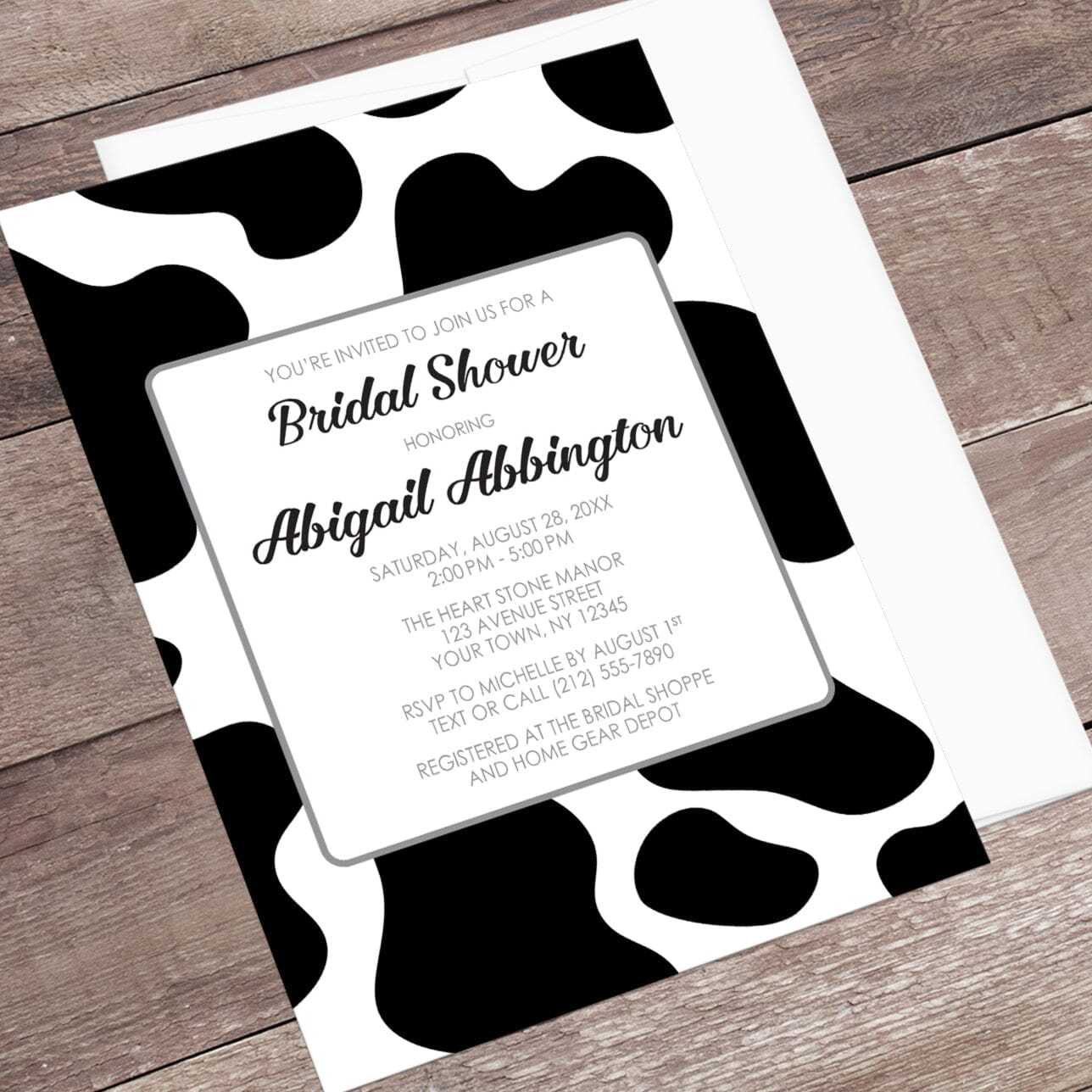 Bridal shower invitation with black and white cow spots pattern, displayed at an angle with a white envelope on a wooden surface