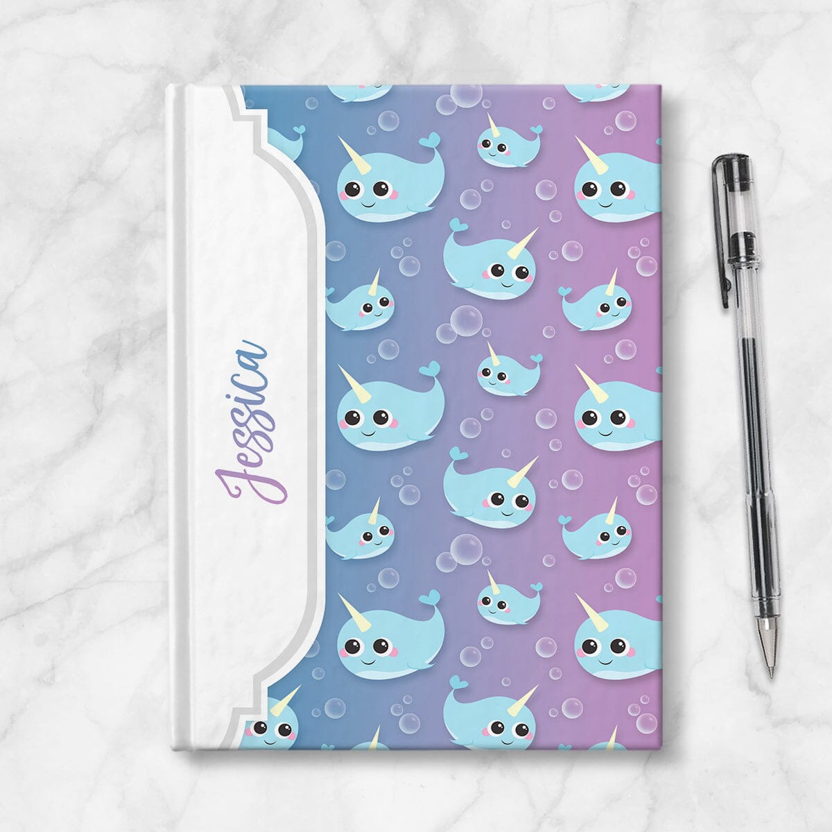 Personalized Cute Happy Narwhal Journal at Artistically Invited. Image shows book on a countertop with a pen next to it.