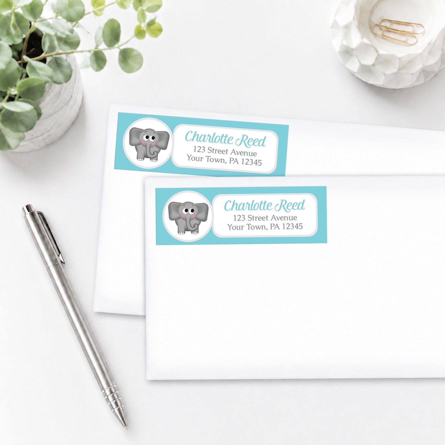 Address labels with a turquoise elephant design on white envelopes, shown on a white surface with a pen and plant.