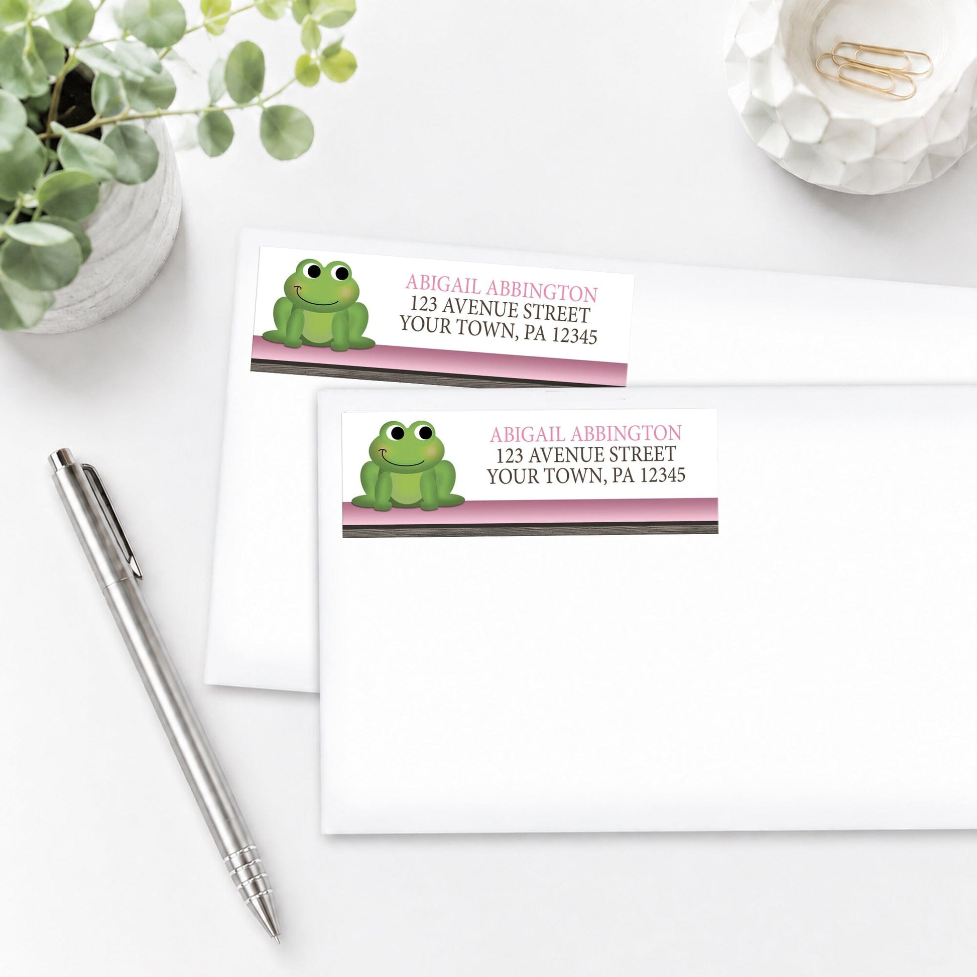 Cute Frog Green Pink Rustic Brown Address Labels, shown on white envelopes, on a white surface with a plant and a pen.
