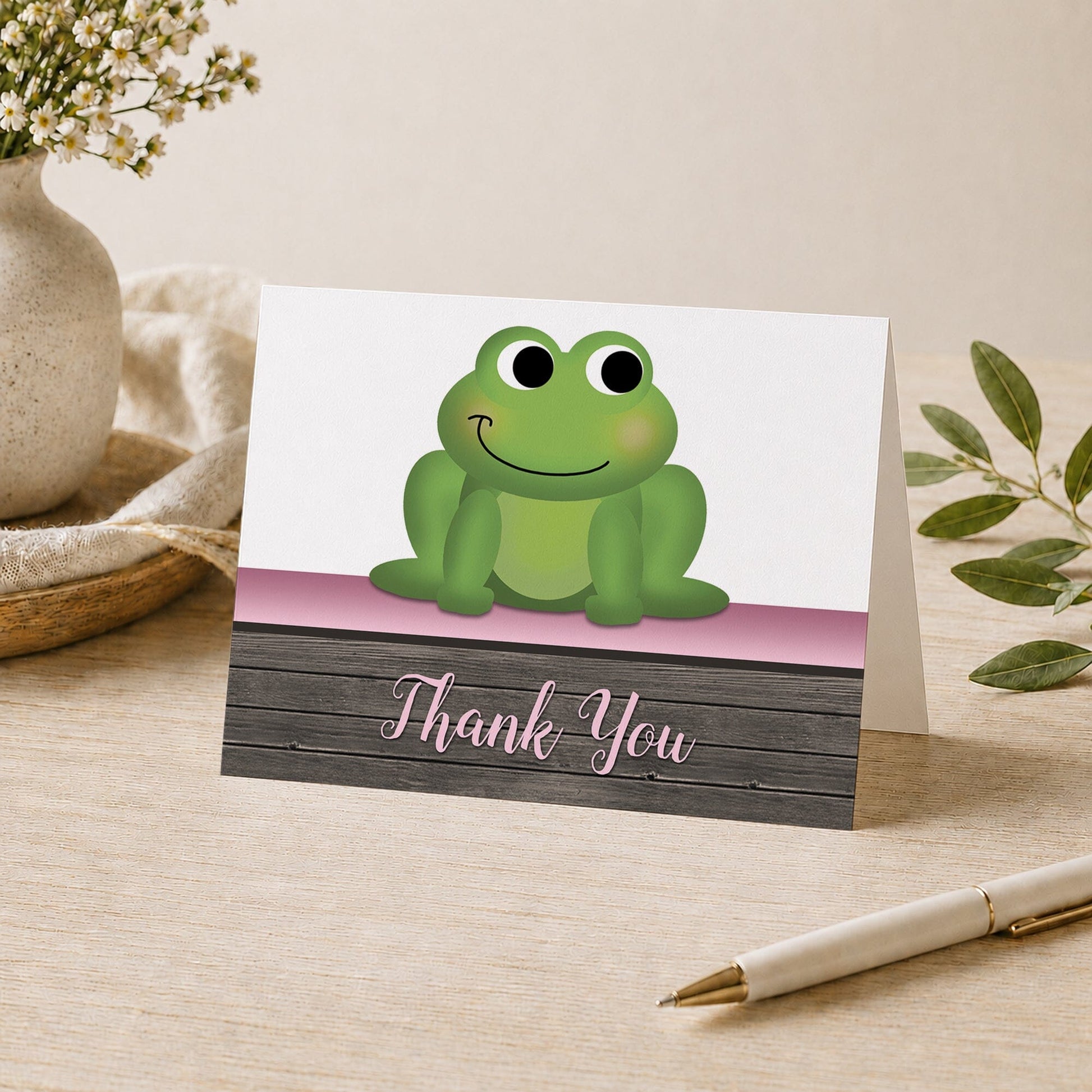 Cute Frog Green Pink Rustic Wood Thank You Card shown on a desk with a vase, plants, and a pen.