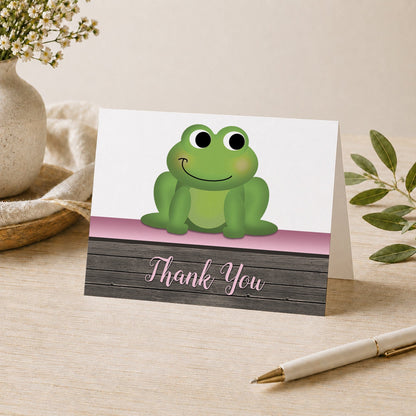 Cute Frog Green Pink Rustic Wood Thank You Card shown on a desk with a vase, plants, and a pen.
