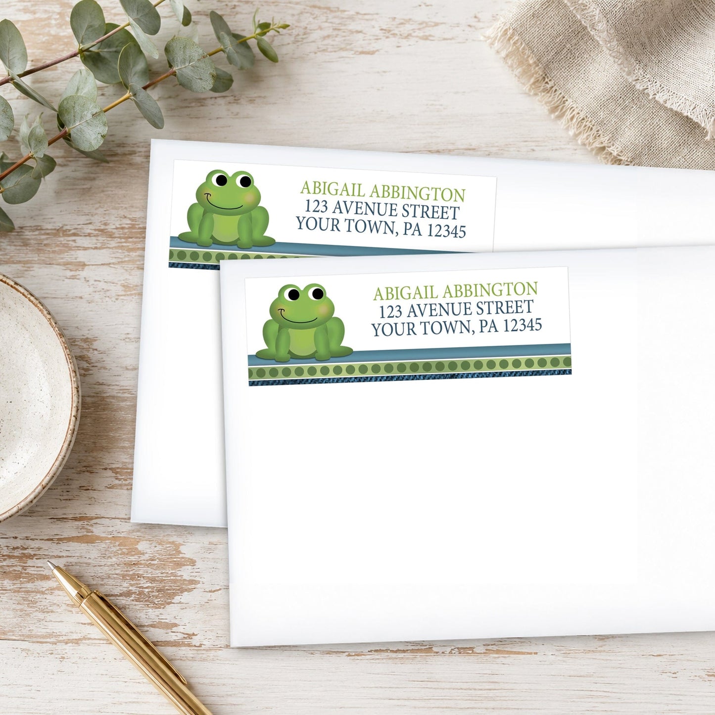 Address labels with frog design on white envelopes on a wooden surface with eucalyptus leaves and a pen.