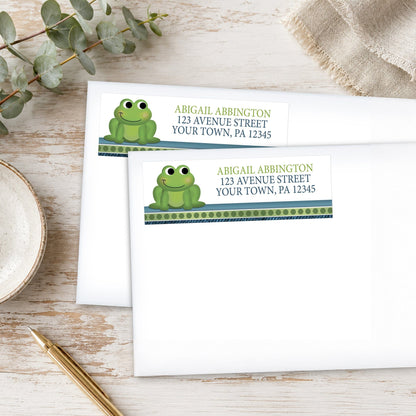 Address labels with frog design on white envelopes on a wooden surface with eucalyptus leaves and a pen.