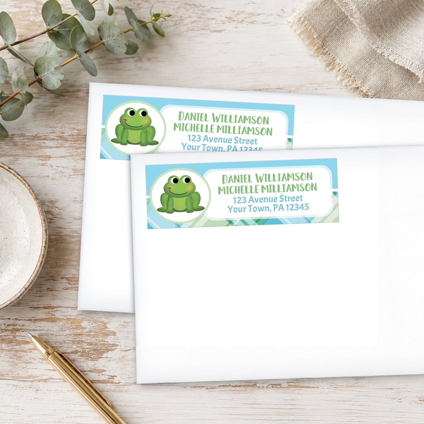 Two address labels with a frog design on a wooden surface with eucalyptus leaves and a pen.
