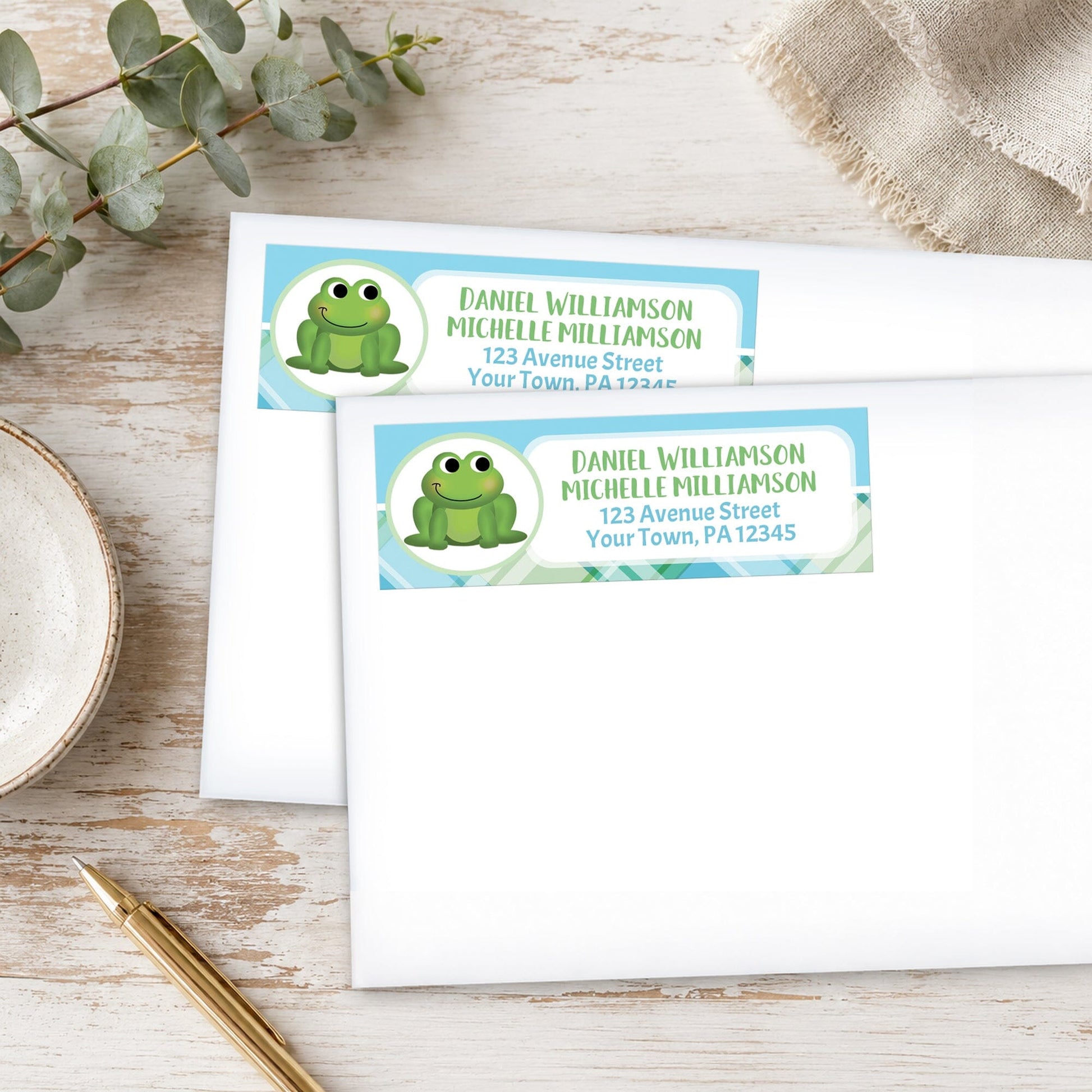 Two address labels with a frog design on a wooden surface with eucalyptus leaves and a pen.