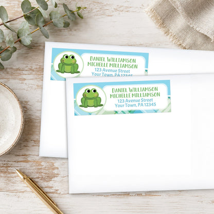 Two address labels with a frog design on a wooden surface with eucalyptus leaves and a pen.
