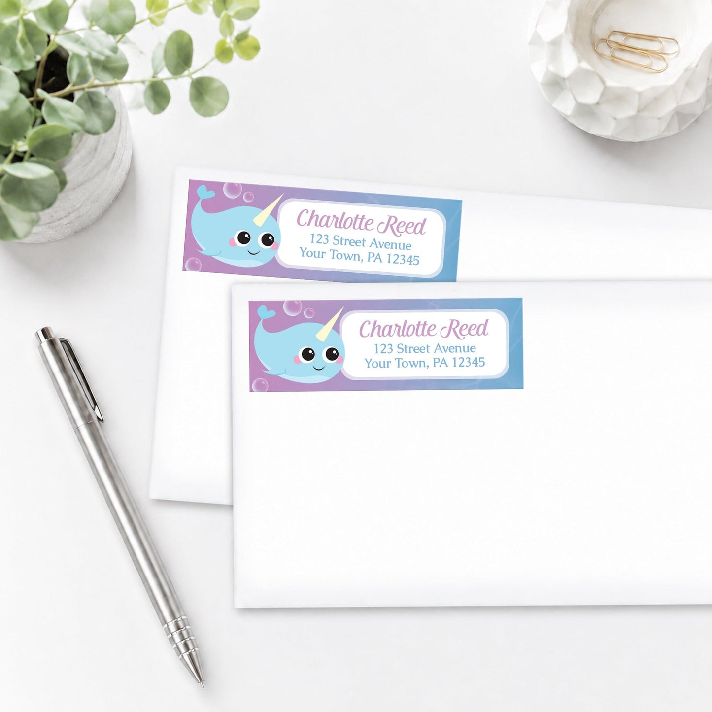 Cute Happy Narwhal Address Labels on white envelopes shown on a white surface with a plant and a pen.